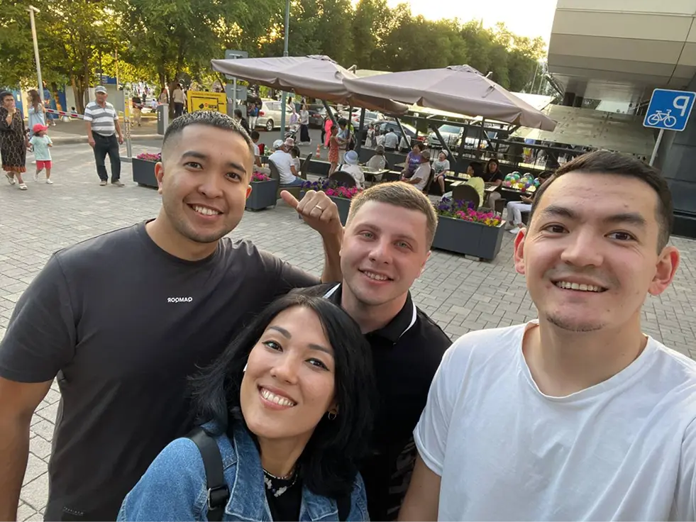 A group of four people smiling and taking a selfie in a lively outdoor setting with tables, umbrellas, and others in the background. Warm, cheerful atmosphere.