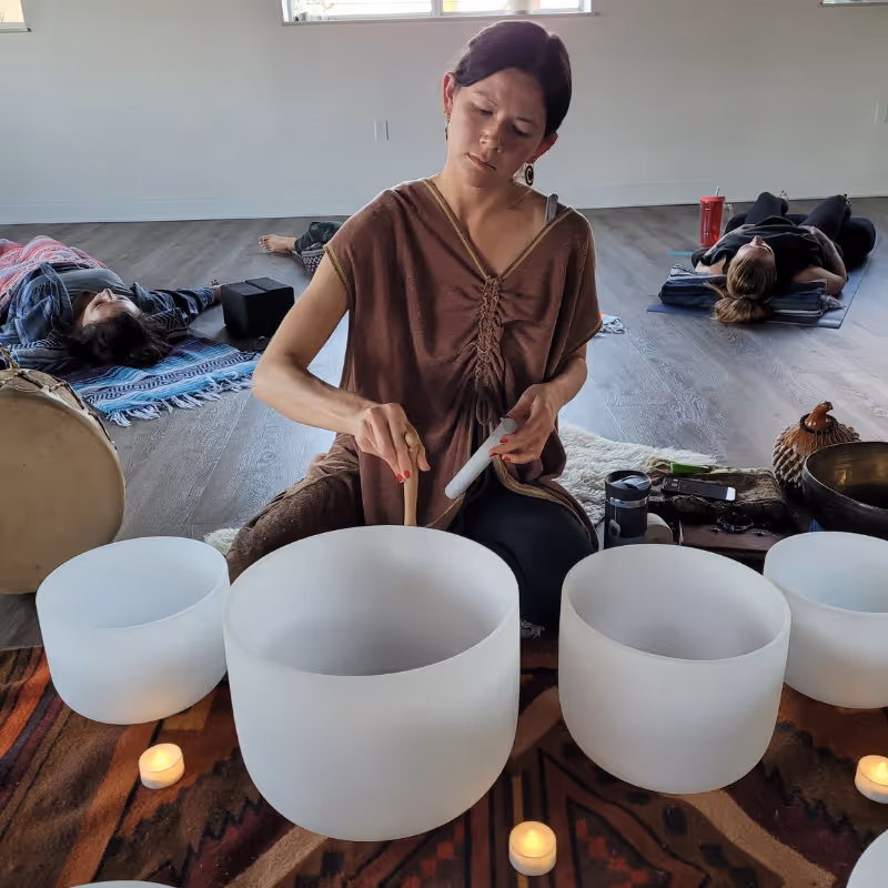 Kayla of All Tree Roots plays crystal singing bowls during a sound bath session, surrounded by candles and participants lying in rest. A peaceful, grounding indoor setting for nervous system support.