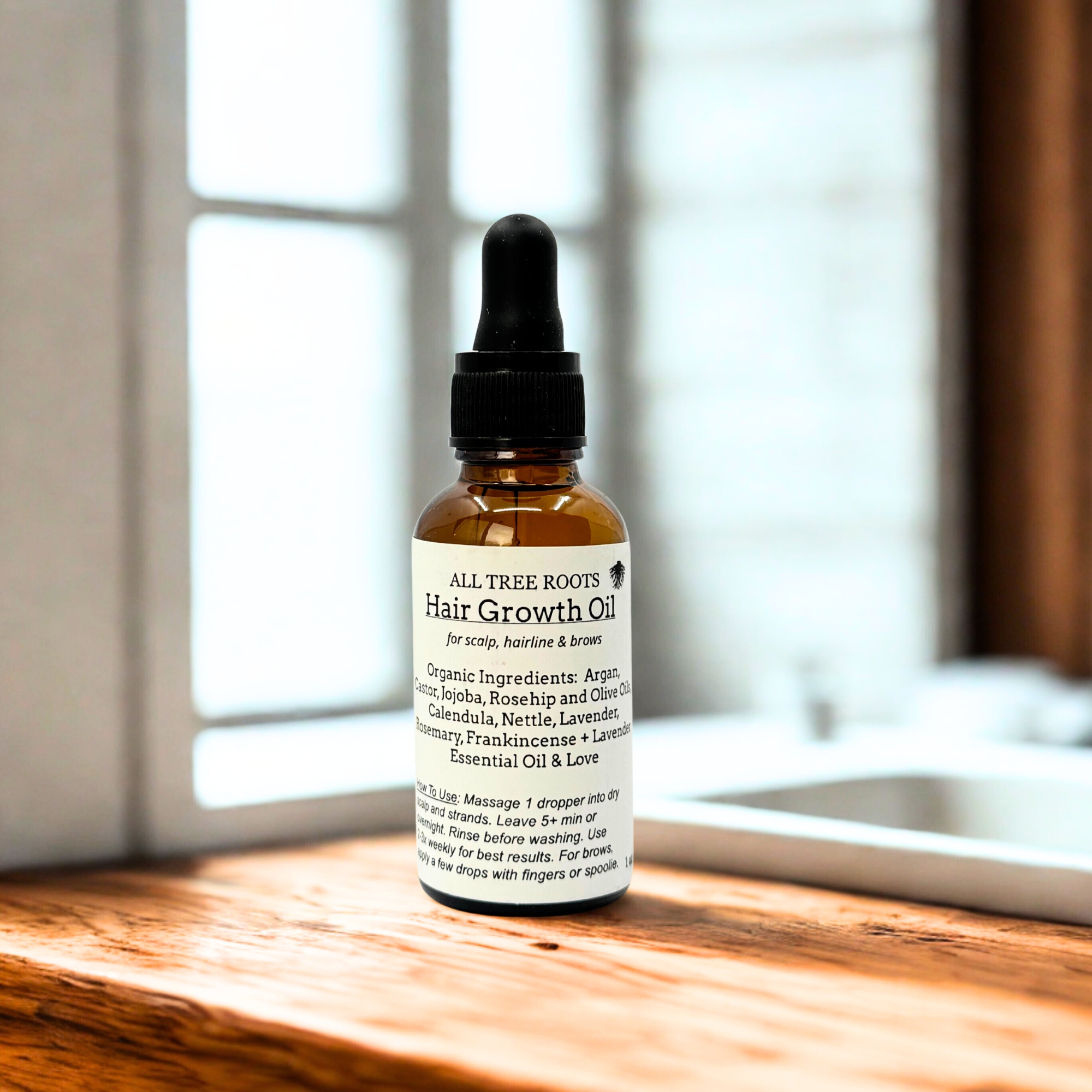 Amber glass dropper bottle of All Tree Roots Hair Growth Oil displayed on a wooden surface in soft natural window light.