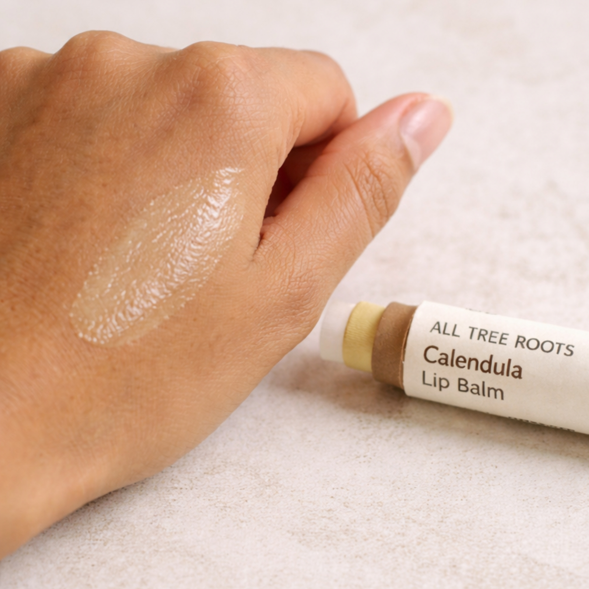 All Tree Roots Calendula Lip Balm in a brown kraft paper tube beside a glossy balm swipe on the back of a hand, styled in soft natural light on a neutral surface.