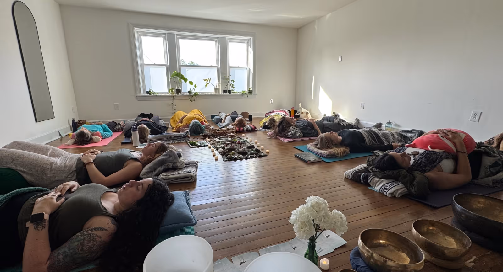 Participants resting on yoga mats during a peaceful group sound bath session in a bright minimalist studio surrounded by singing bowls, candles, and natural light.