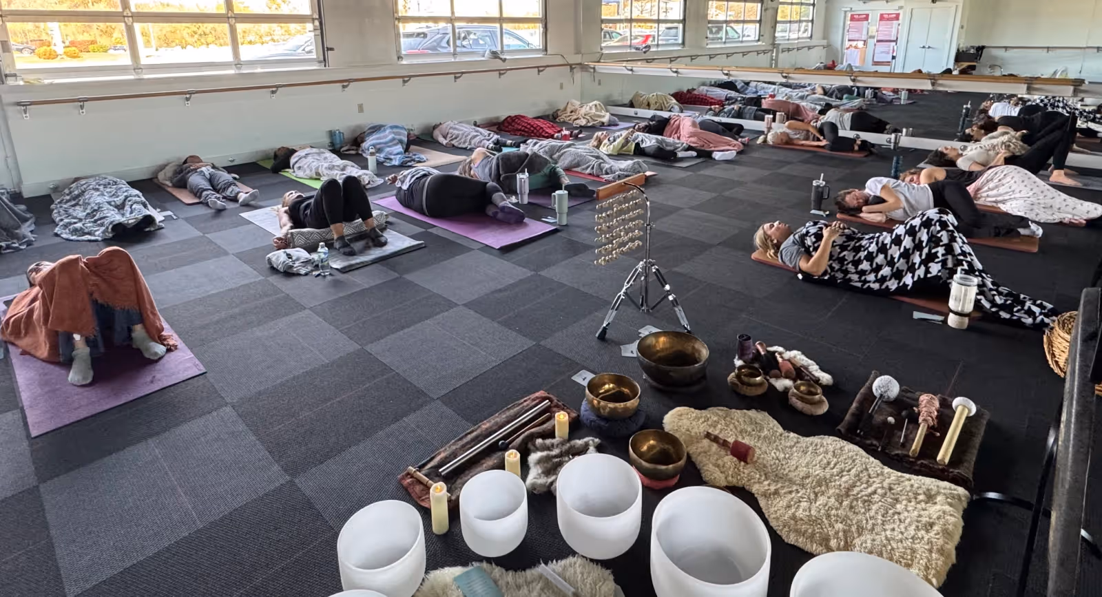 Participants resting on yoga mats during a large group sound bath session surrounded by crystal bowls, singing bowls, and calming instruments in a bright studio space.
