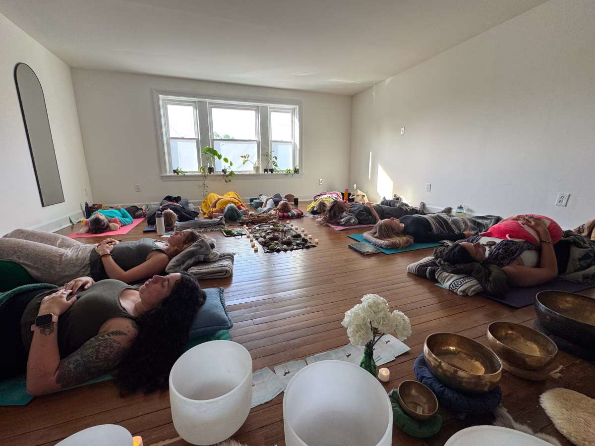 Participants rest on yoga mats under blankets during a sound bath, with crystal and metal singing bowls in the foreground of a bright studio.