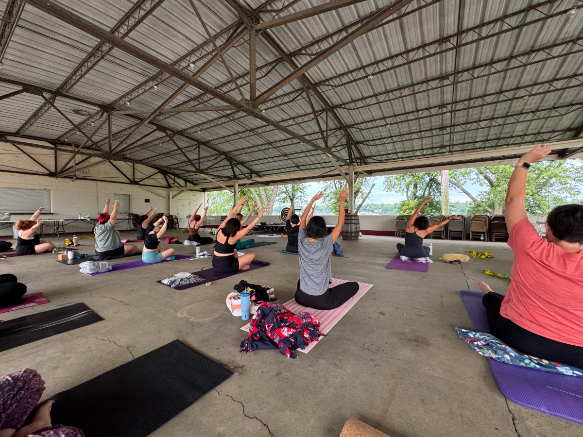 A group practices seated yoga with arms raised on colorful mats beneath an open-sided outdoor pavilion.