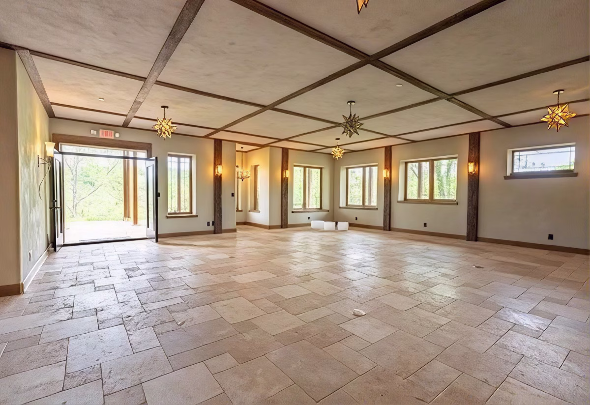 An empty studio features stone tile flooring, exposed wooden beams, star-shaped pendant lights, and windows overlooking greenery.