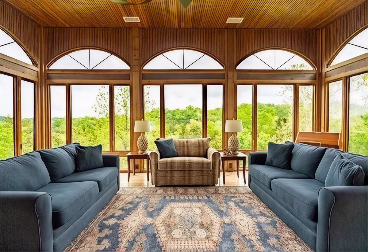 Two blue sofas and a plaid armchair surround a patterned rug in a wood-paneled sunroom with tall arched windows overlooking green trees.