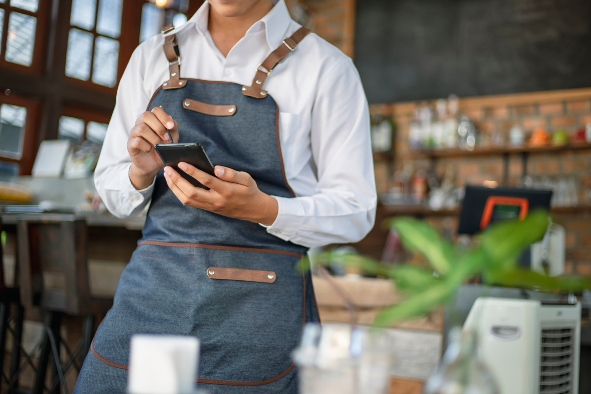 Restaurant staff member using a smartphone behind the counter near a point-of-sale terminal.