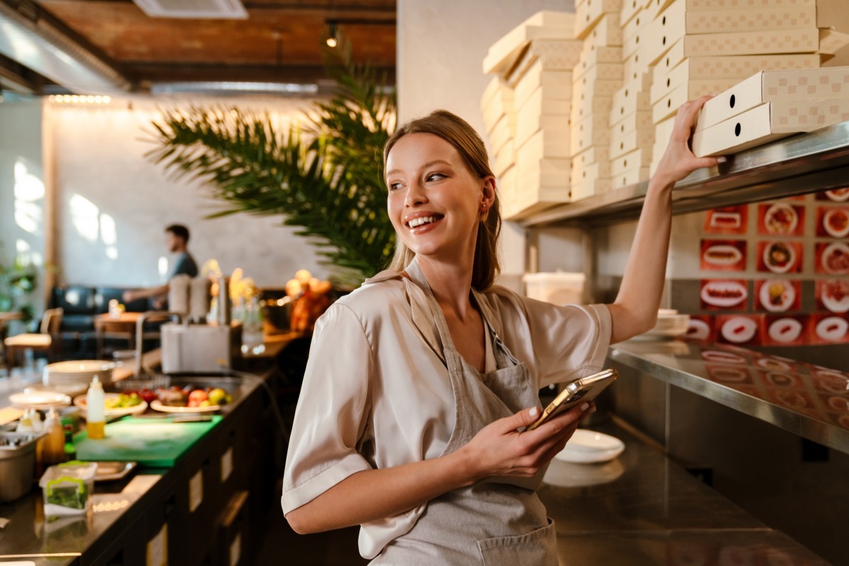 Restaurant employee holding a smartphone while reaching for a takeout box in a busy kitchen.