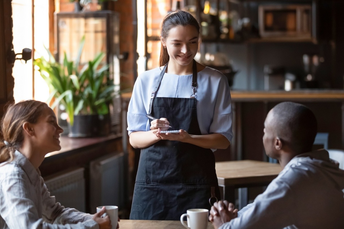 Restaurant server writing down an order while speaking with two guests at their table.