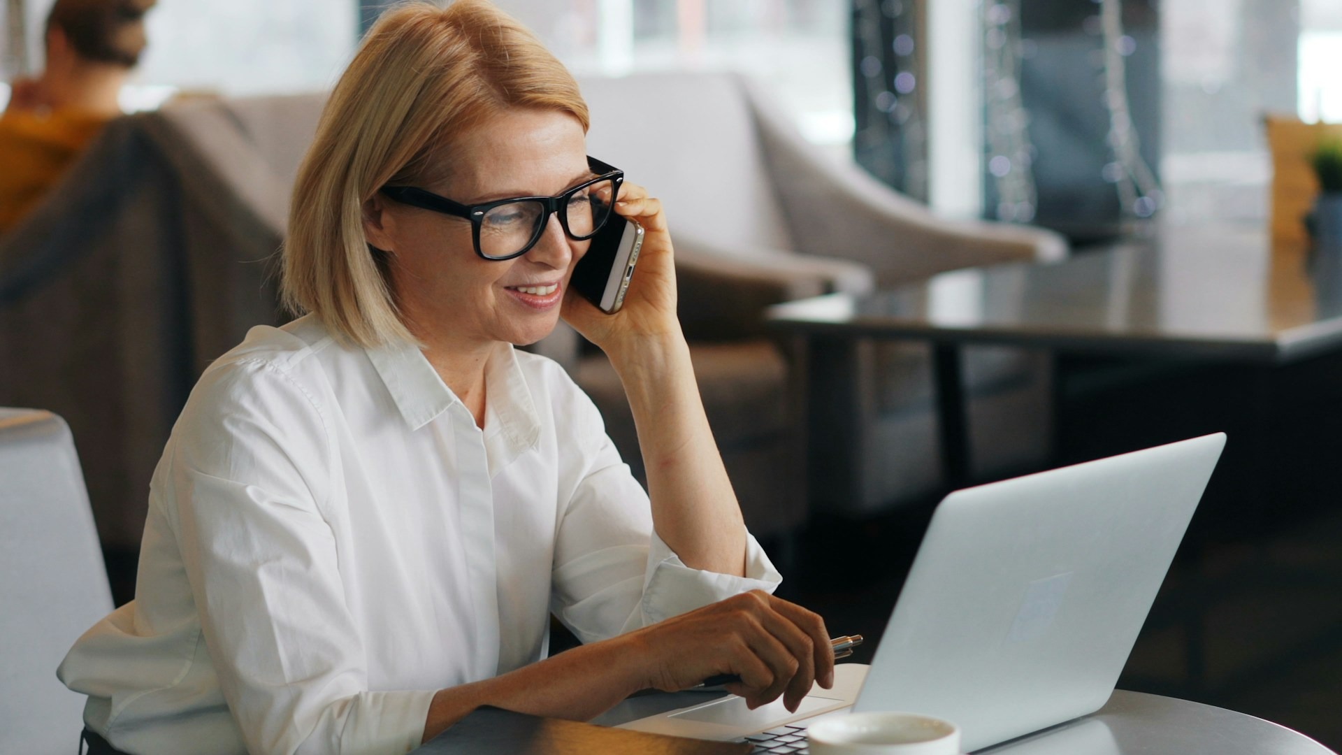A businesswoman wearing glasses smiles while taking a call on her smartphone, one hand resting on her laptop keyboard, with a coffee cup nearby in a softly lit café.