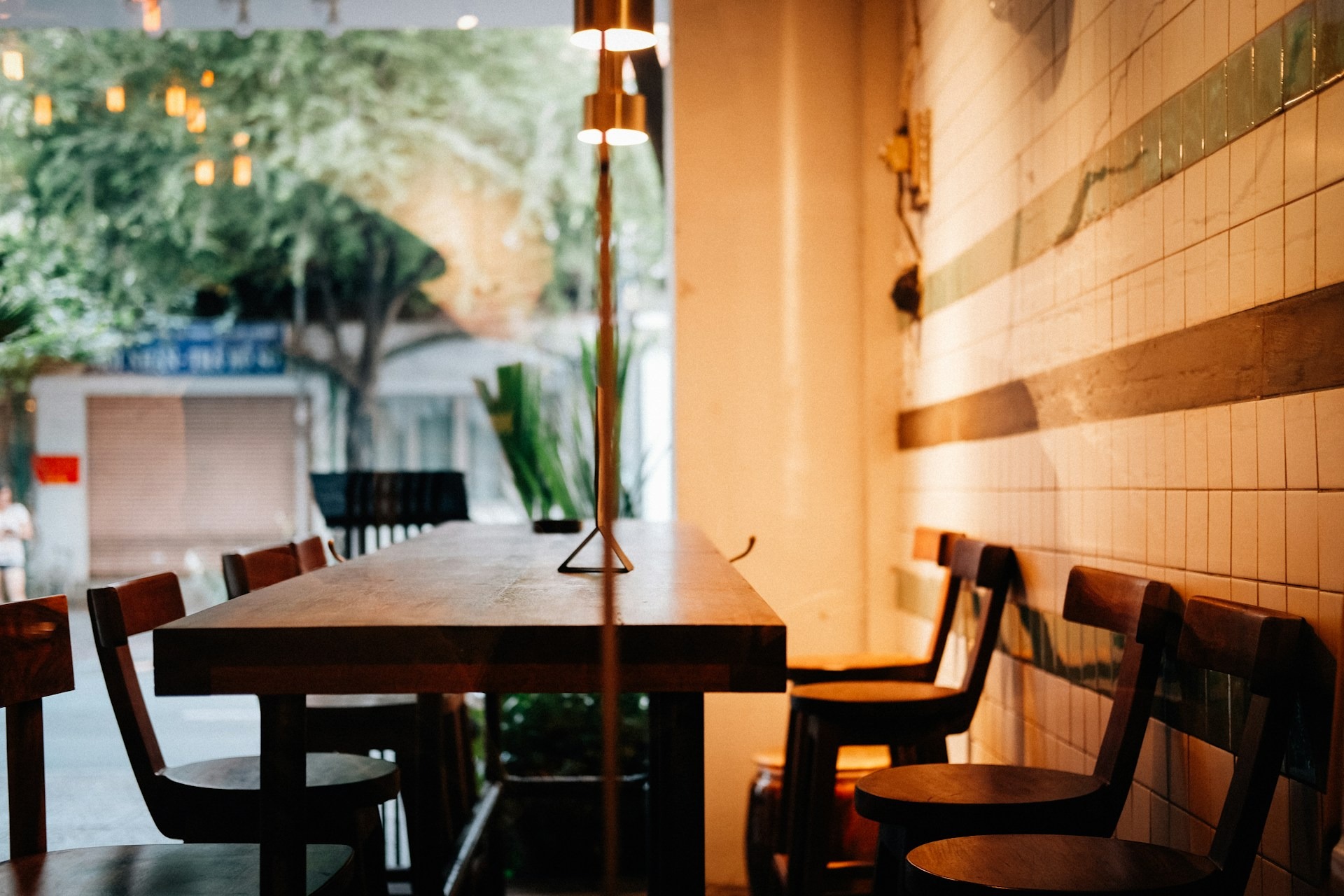 A quiet dining nook with a dark wood table, mismatched wooden chairs, tiled wall, and a brass floor lamp, with string lights and greenery visible in the background