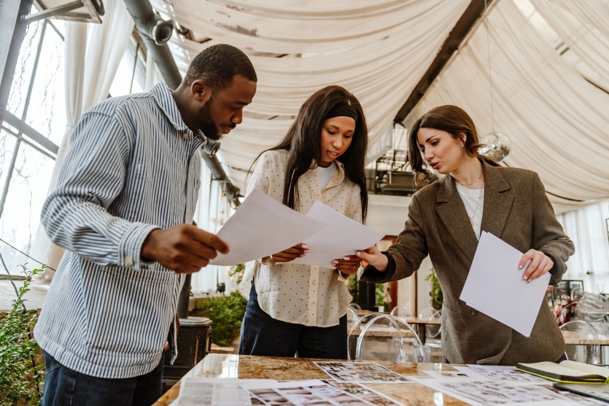 Three colleagues standing at a table in an event space, comparing printed pages and pointing out details on one of them.