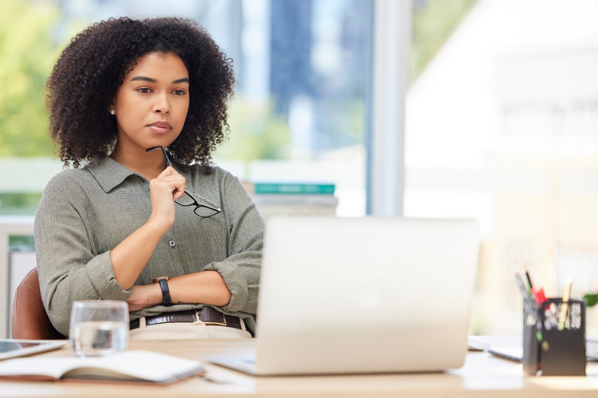 Woman at a laptop holding her glasses, looking thoughtful while reviewing something on screen at a desk with a notebook and water glass.