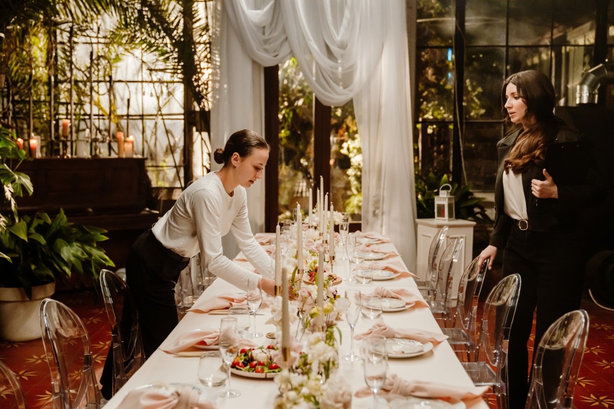 Venue staff setting a long formal dining table with pink linens, candlesticks, and floral centerpieces inside a draped event venue.