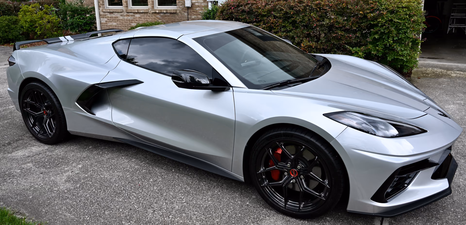 Silver C8 Corvette with Forgelite SV00 aftermarket wheels car with black rims and red brake calipers parked on a driveway in front of a house with brick wall and greenery.