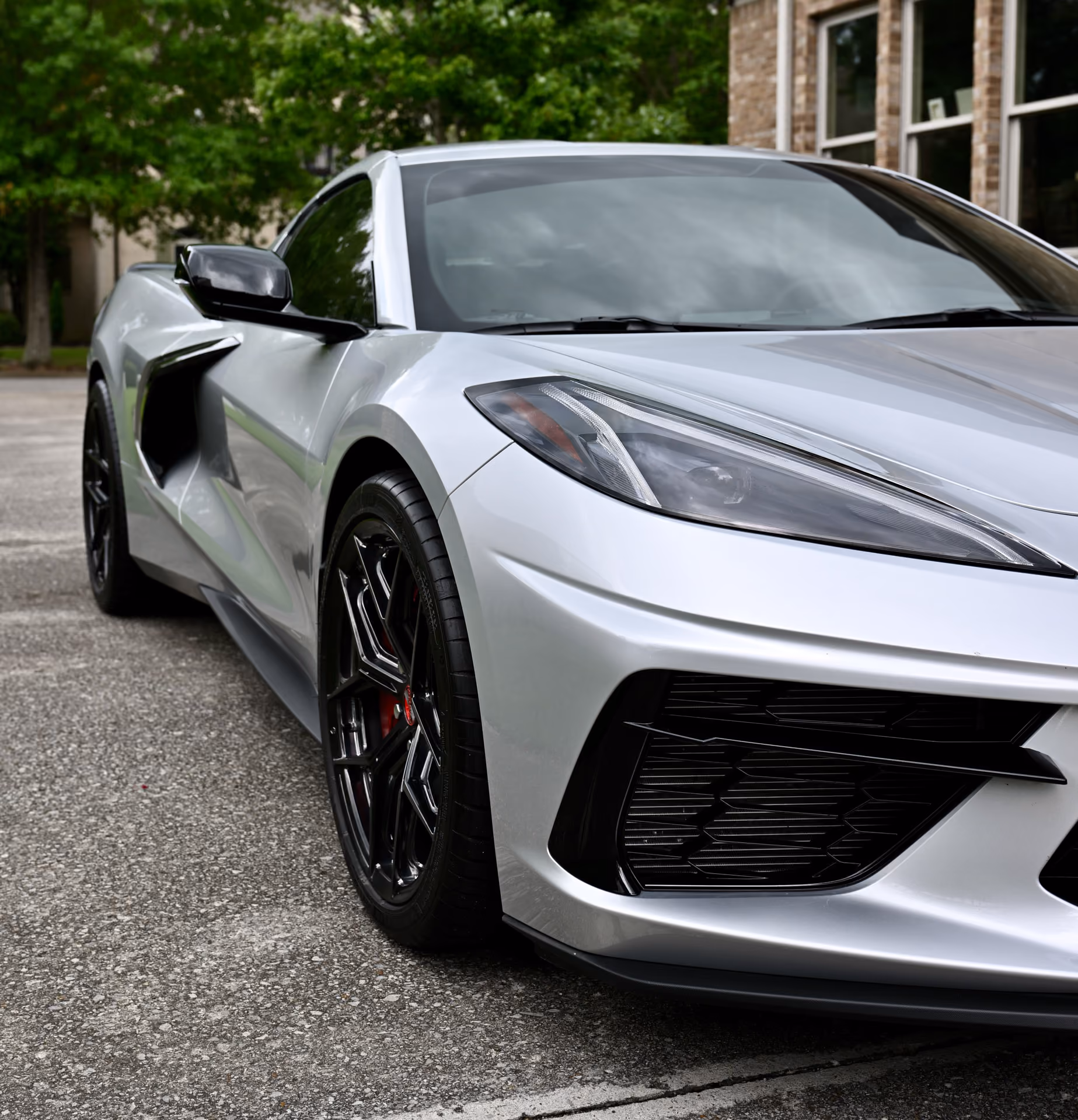 Close-up view of the front left side of a silver C8 Corvette with Forgelite SV00 aftermarket wheels, parked on a paved surface with greenery and a brick building in the background.