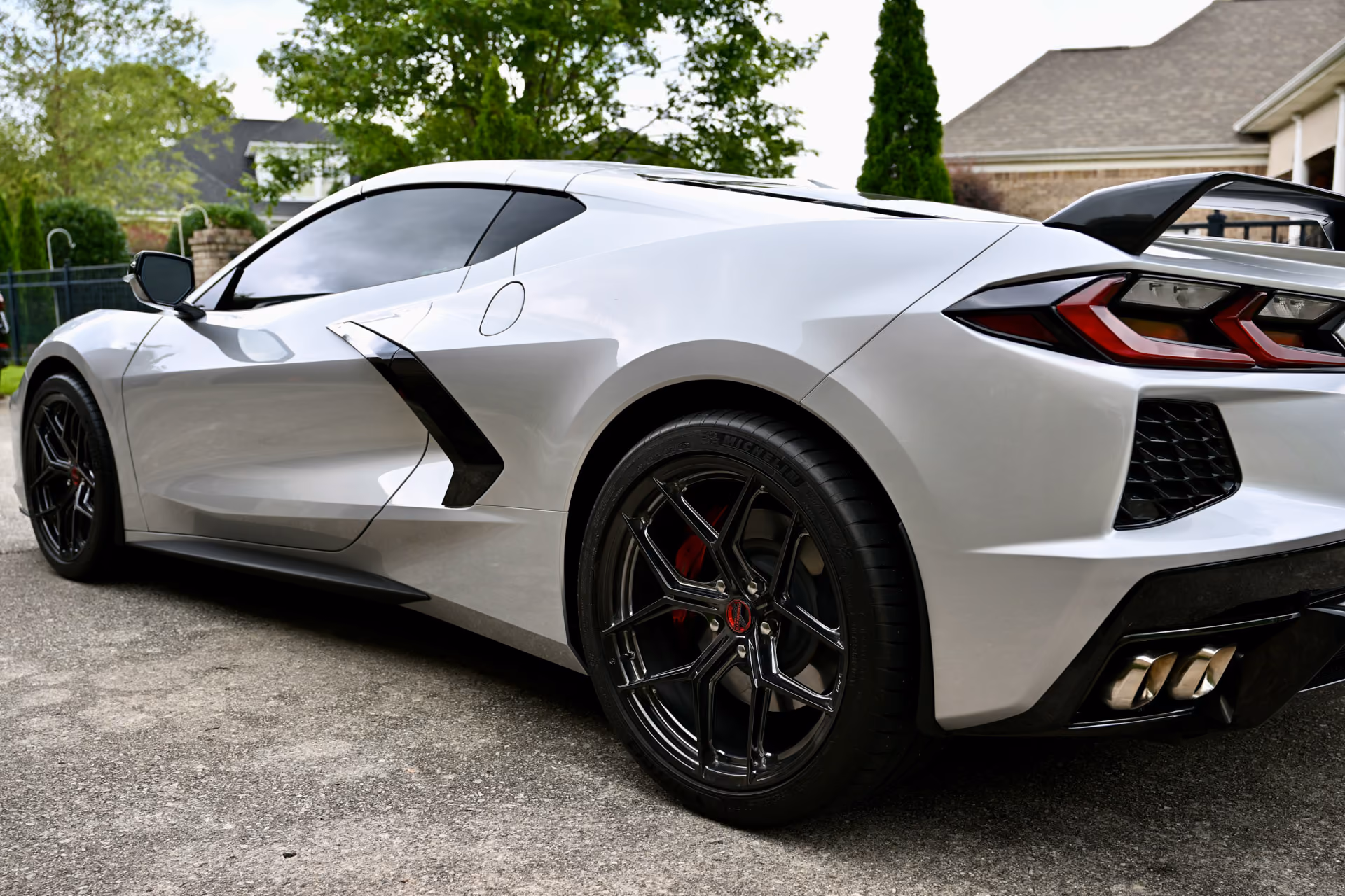 Silver C8 Corvette with Forgelite SV00 aftermarket wheels parked on a driveway near suburban houses and green trees, viewed from the rear side showing black wheels, rear spoiler, and quad exhaust pipes.