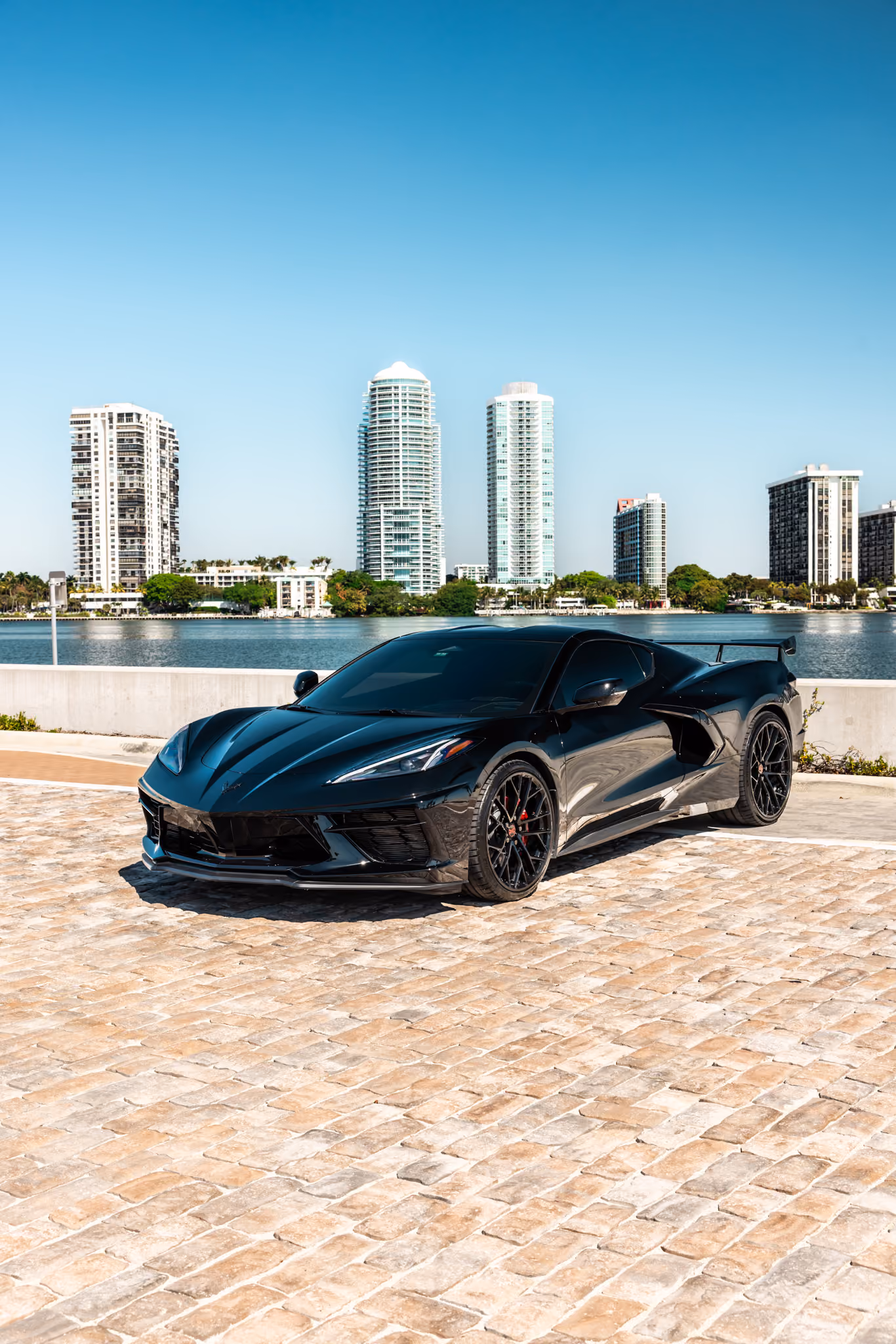 Black C8 Corvette with Forgelite LP4 aftermarket wheels parked on a cobblestone surface with a body of water and several tall buildings in the background under a clear blue sky.
