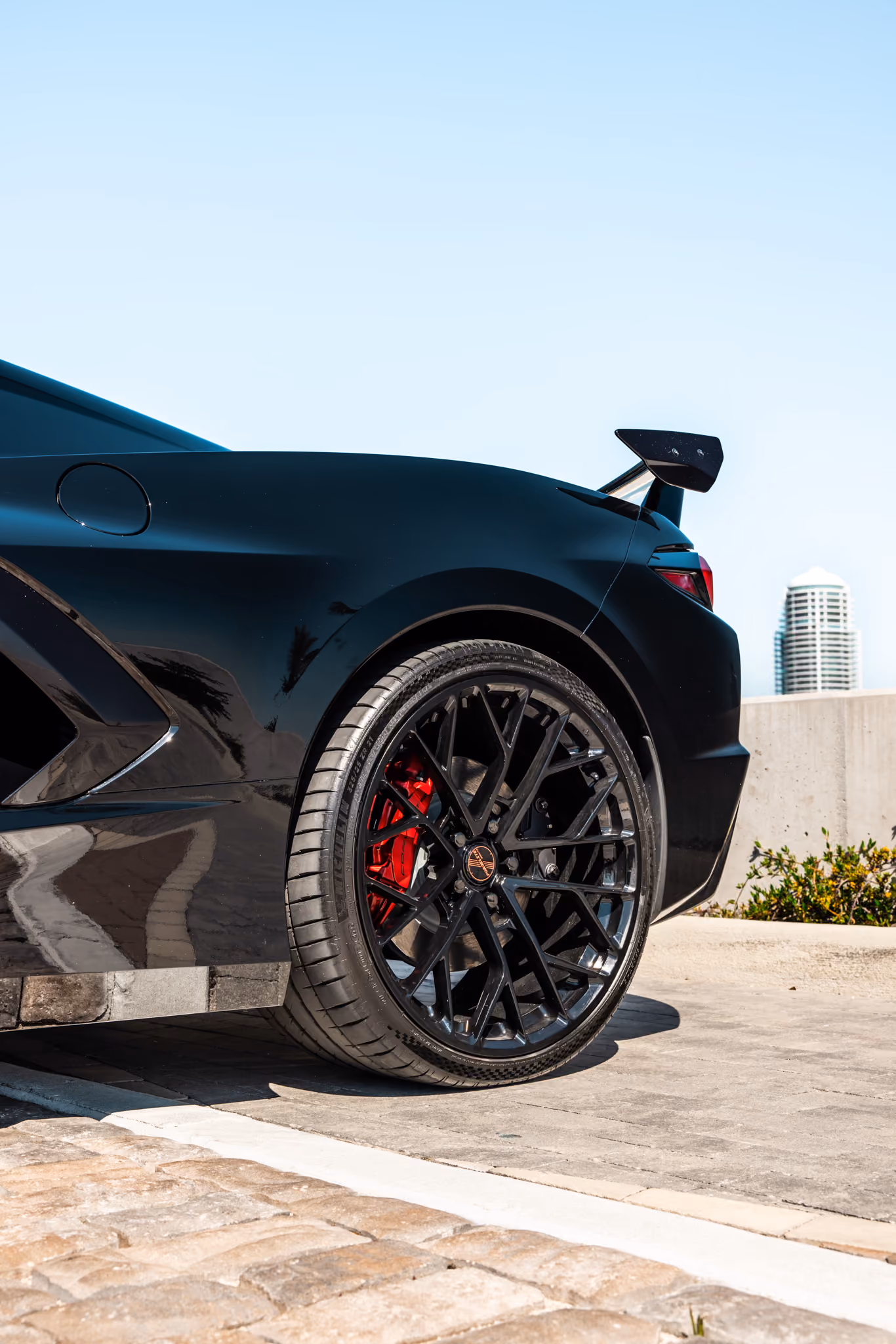 Rear side view of a C8 Corvette with Forgelite LP4 aftermarket wheels parked outdoors showing a stylish large black alloy wheel with a red brake caliper and a raised rear spoiler, under clear blue sky.