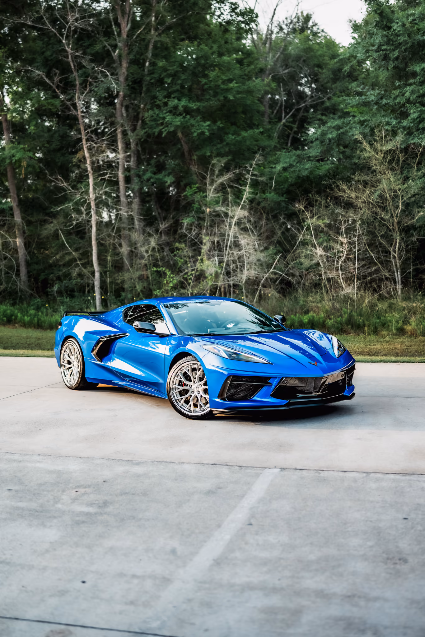 Bright blue C8 Corvette with Forgelite FL4-M parked on a concrete surface with green trees and foliage in the background.