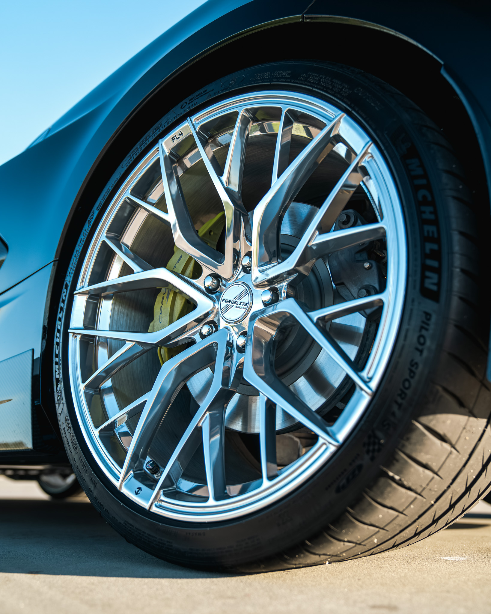 Close-up of a shiny multi-spoke Forgelite FL4 alloy wheel mounted with a Michelin Pilot Sport tire on a sports car, showing a yellow brake caliper and ventilated brake disc.