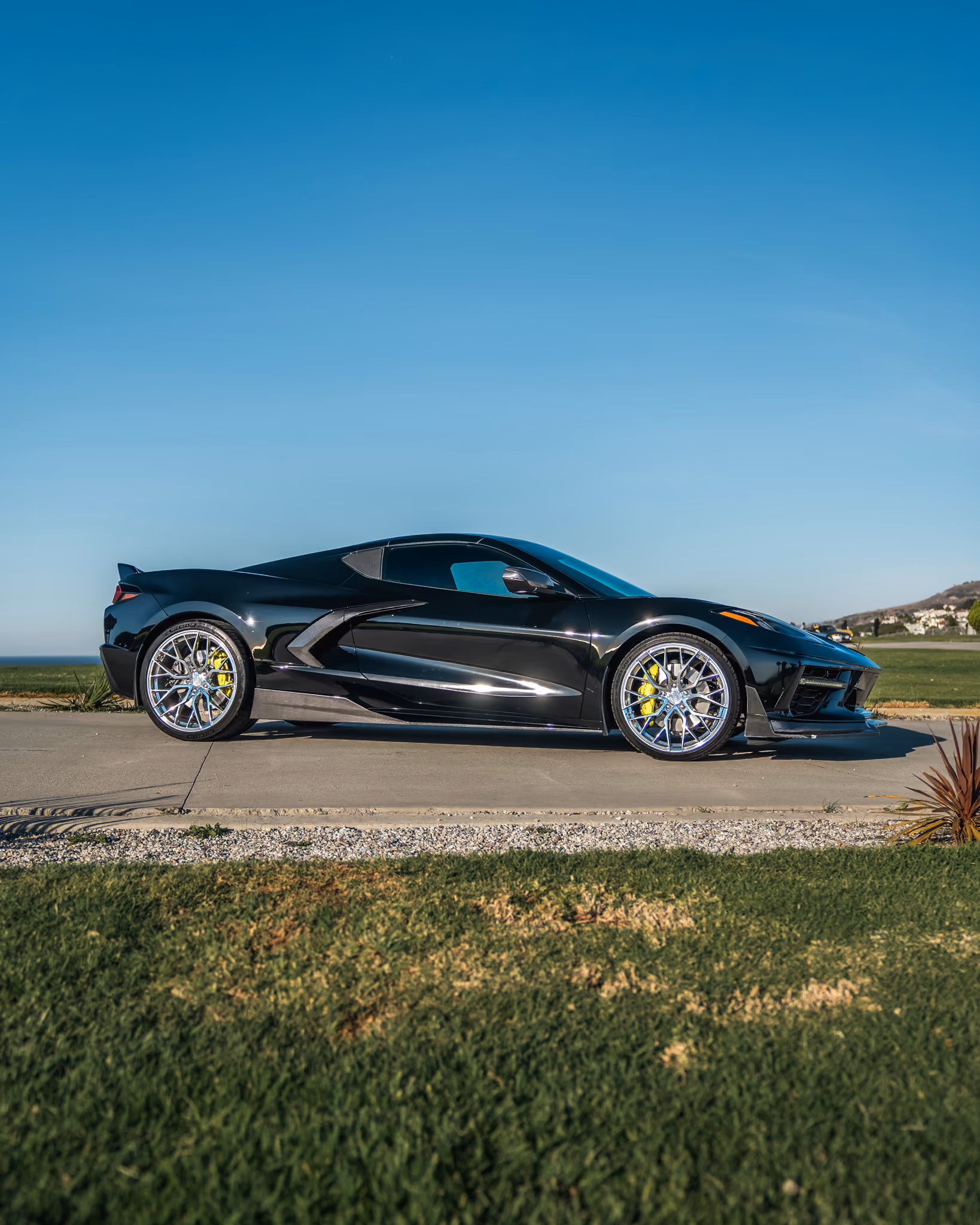 Sleek black C8 Corvette with Forgelite FL4 parked on a concrete path with green grass in the foreground and a clear blue sky in the background.