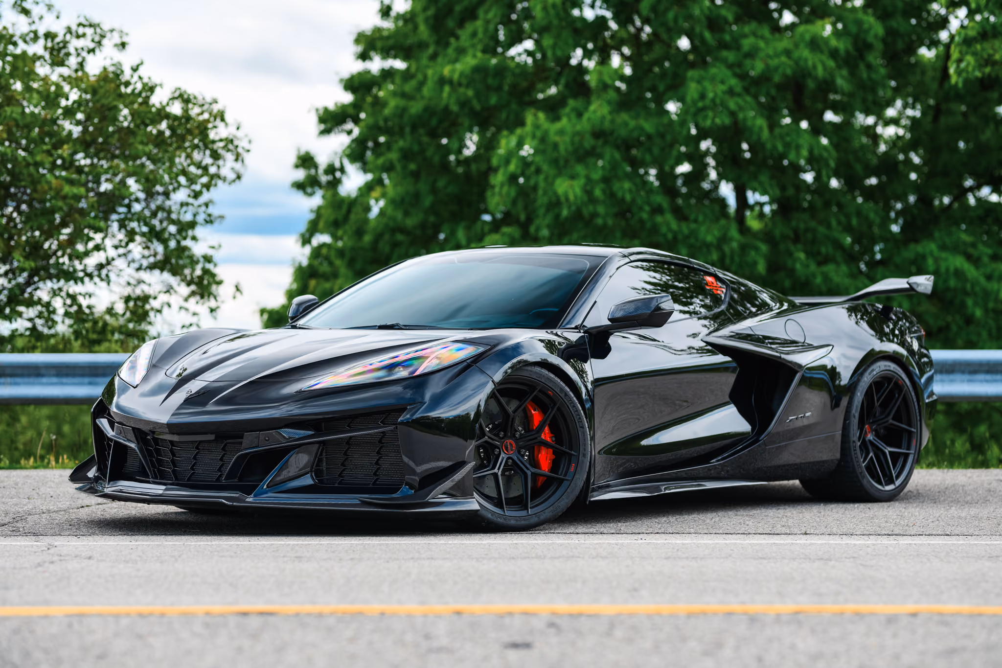 Black Corvette C8 Z06 with Forgelite SV00 aftermarket wheels, with red brake calipers, parked on asphalt road with green leafy trees in the background.