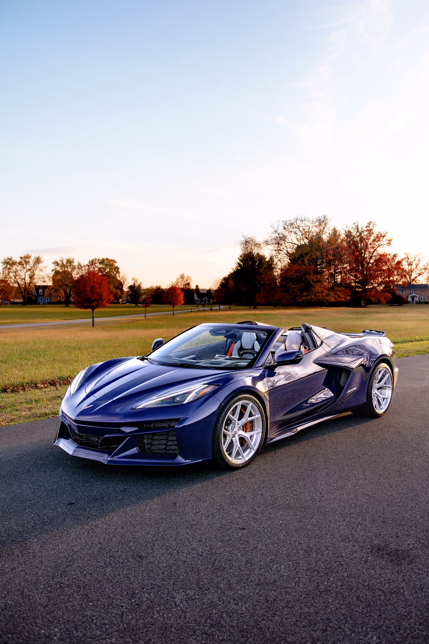 Purple Corvette C8 with Forgelite FL1 aftermarket wheels parked on a paved road beside a grassy field with autumn trees and houses in the background during sunset.