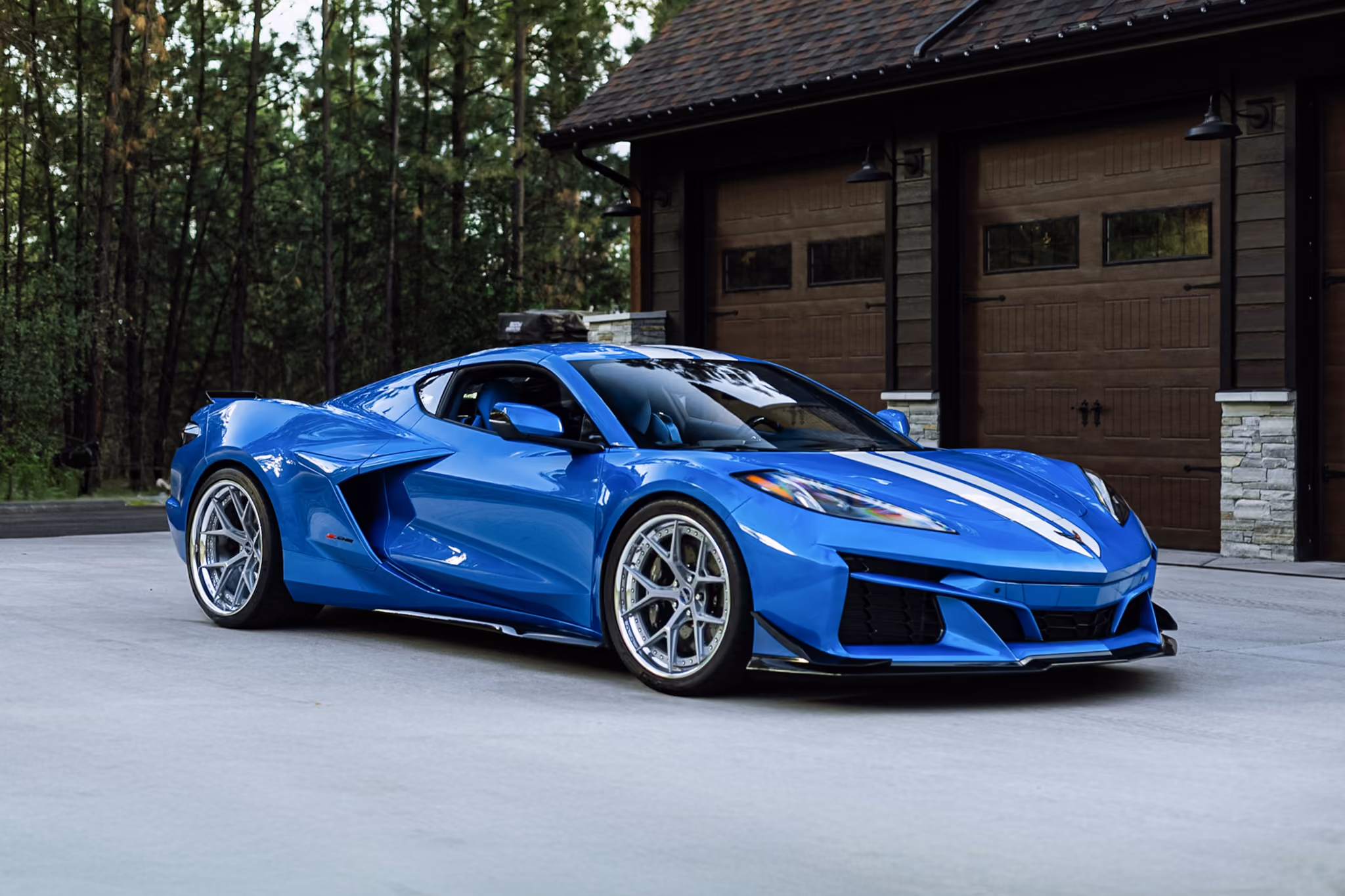 Blue Chevrolet Corvette with Forgelite FL1-M aftermarket wheels with white racing stripes parked on a concrete driveway in front of a wooden garage with stone pillars, surrounded by tall trees.
