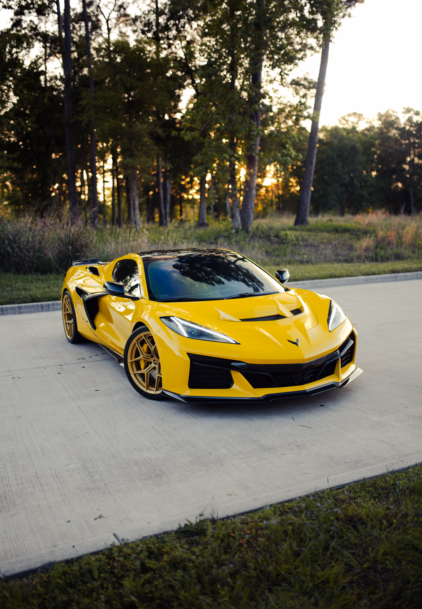 Yellow Chevrolet Corvette C8 sports car with Forgelite SV00 aftermarket wheels black roof and gold wheels parked on a concrete road with trees and grass in the background at sunset.
