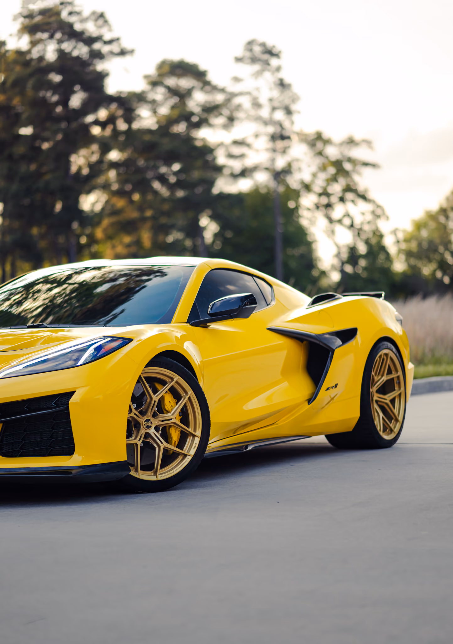 Yellow Chevrolet Corvette C8 ZR1 sports car with gold Forgelite wheels SV00 parked on a concrete surface with trees in the background.