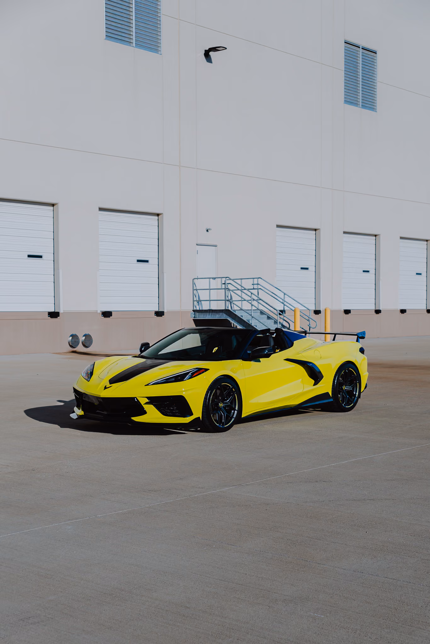 Bright yellow Chevrolet Corvette C8 with Forgelite SV00 aftermarket wheels, with black accents parked in front of a white industrial building with multiple closed garage doors and a metal staircase.