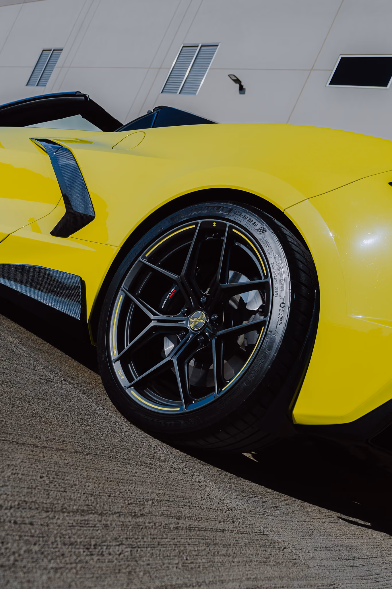 Close-up of the rear side of a Chevrolet Corvette C8 with Forgelite SV00 aftermarket wheels with a silver rim accent, parked on a textured concrete surface against a background of a beige building with windows and a light fixture.