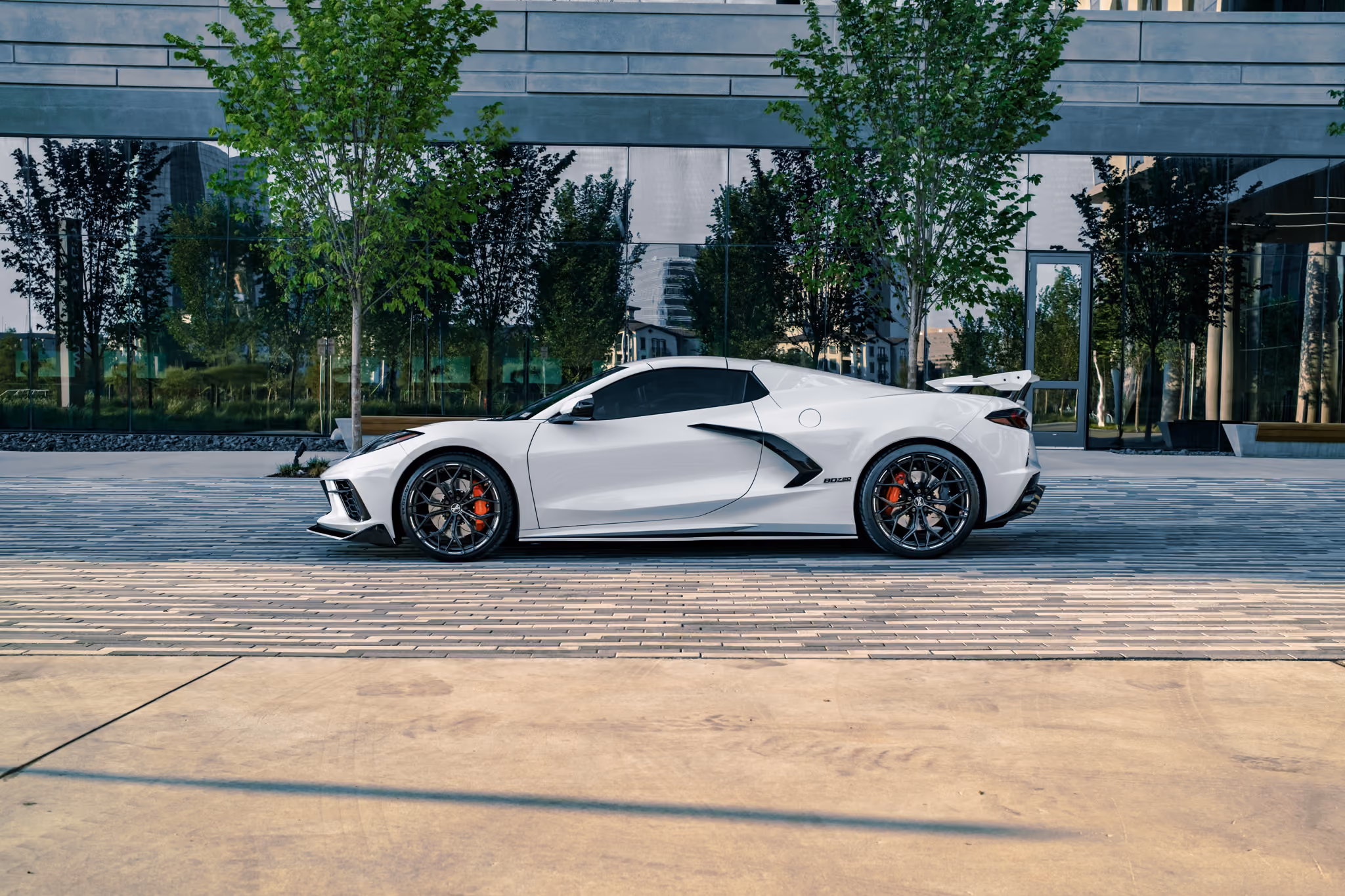 Side view of a Corvette C8 with Forgelite SV10 aftermarket wheels and red brake calipers parked on a patterned paved surface in front of a modern building with large reflective windows and green trees.