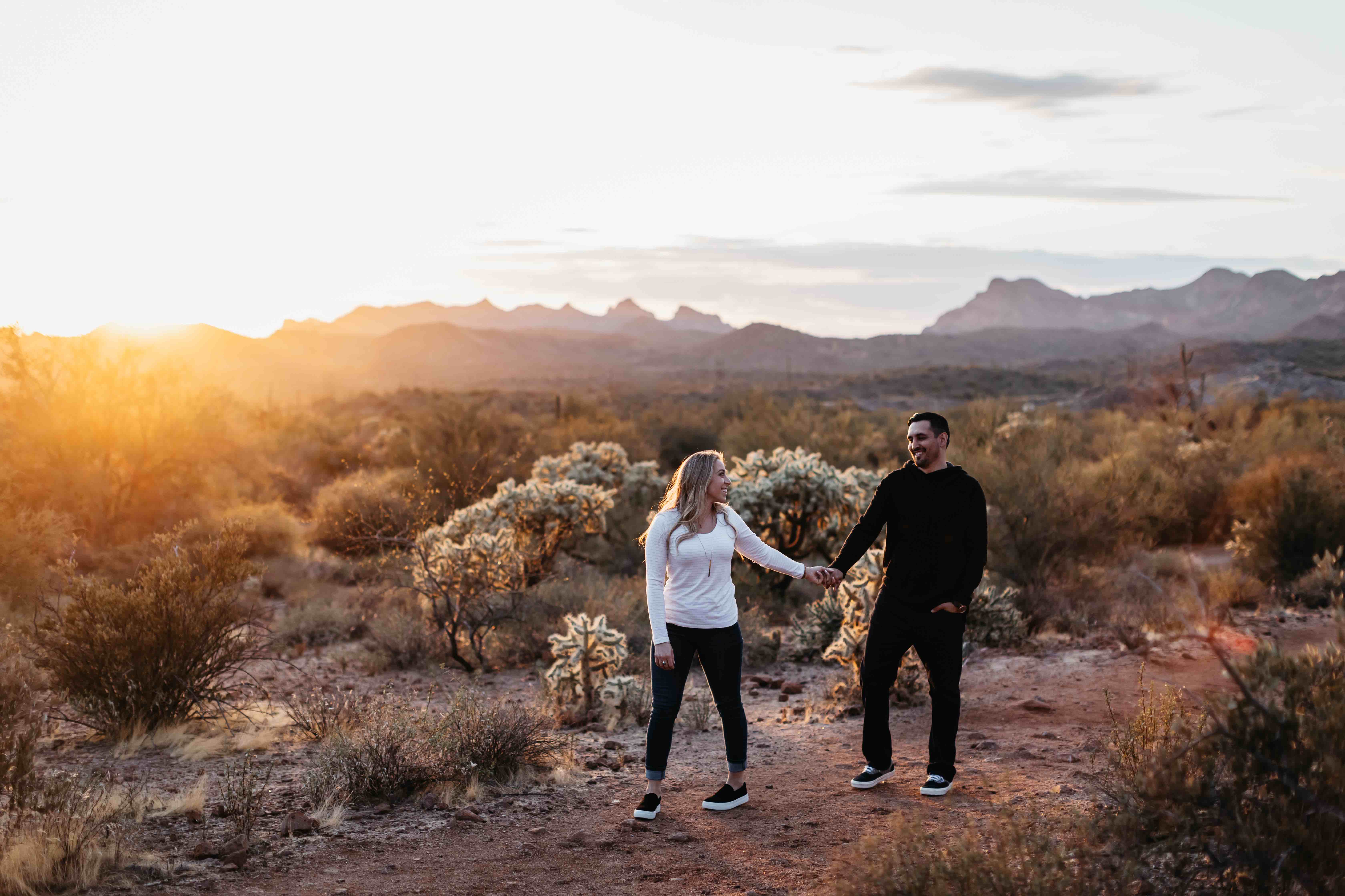 Couple holding hands and smiling while standing on a desert path at sunset with mountains behind.