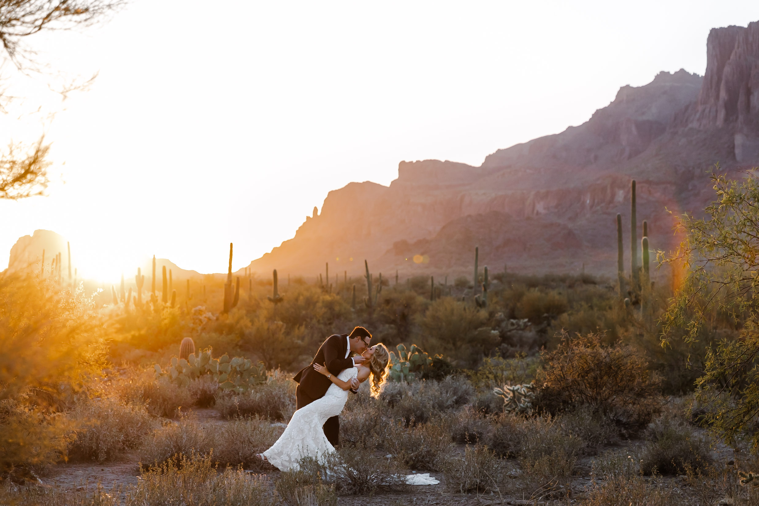 Bride and groom kissing in Arizona desert at sunset with mountains and cacti in the background.