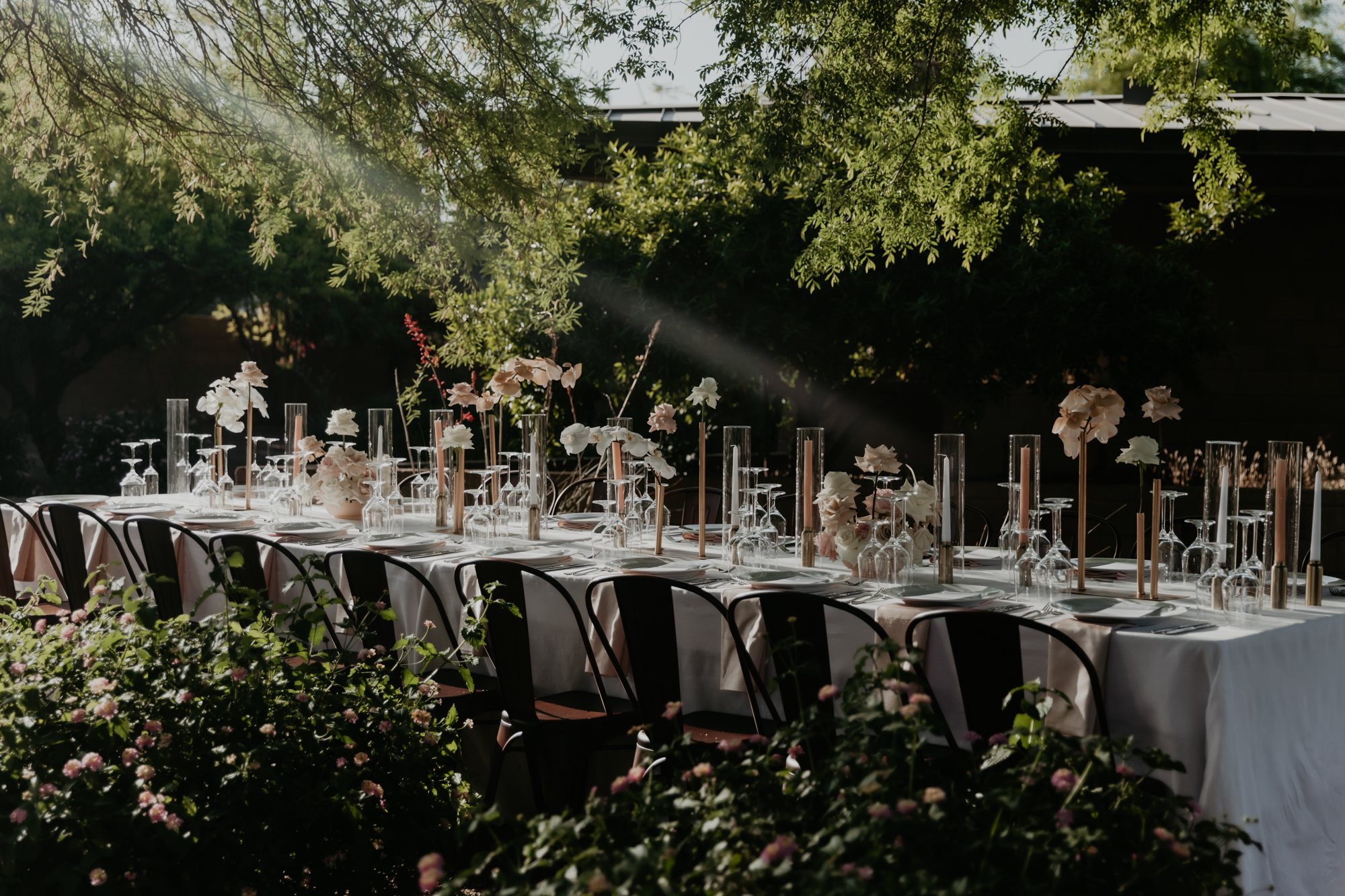 Outdoor long dining table set with white tablecloth, flowers, candles, and upside-down wine glasses.