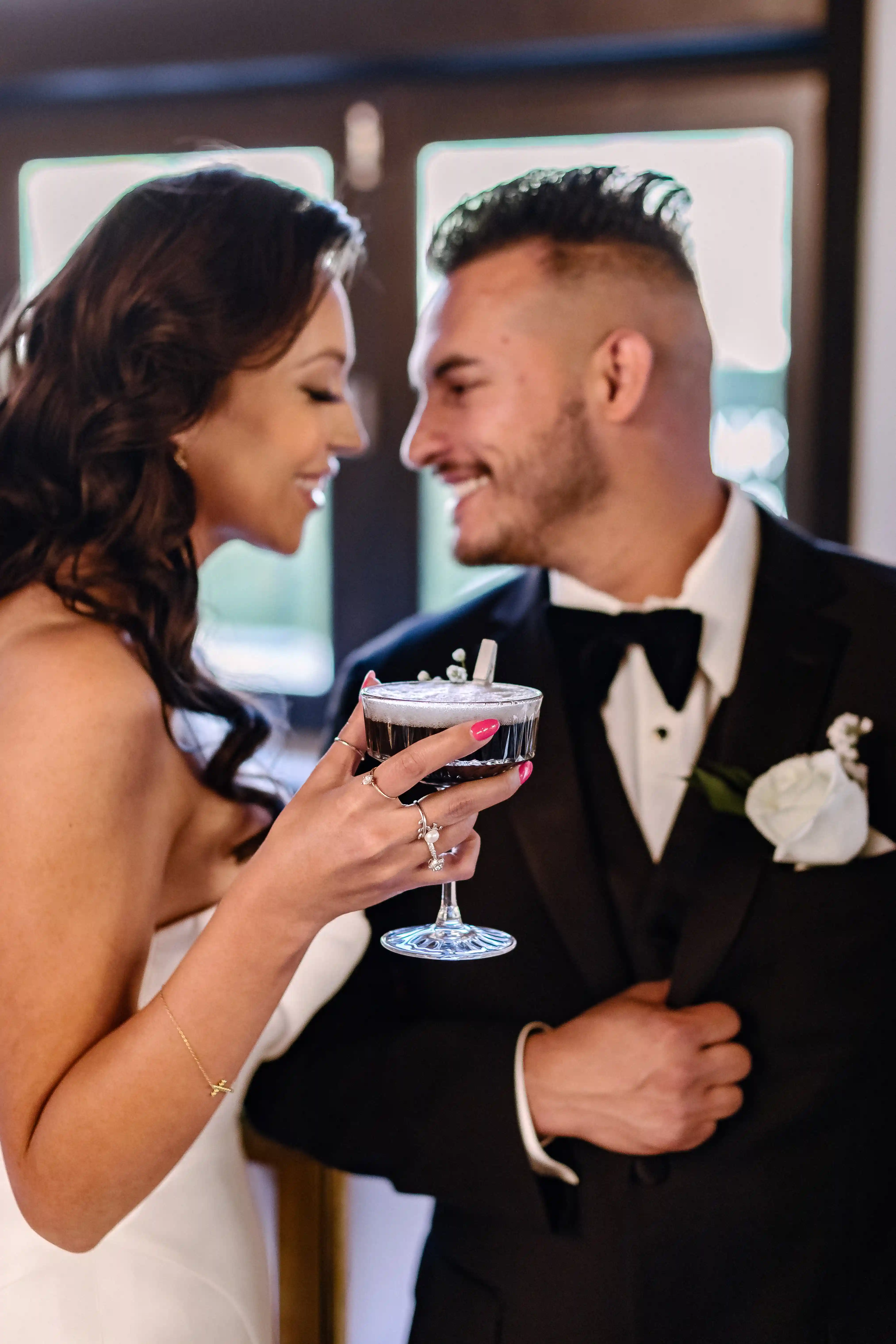 Bride and groom smiling at each other, bride holding a cocktail glass with dark drink and garnish.