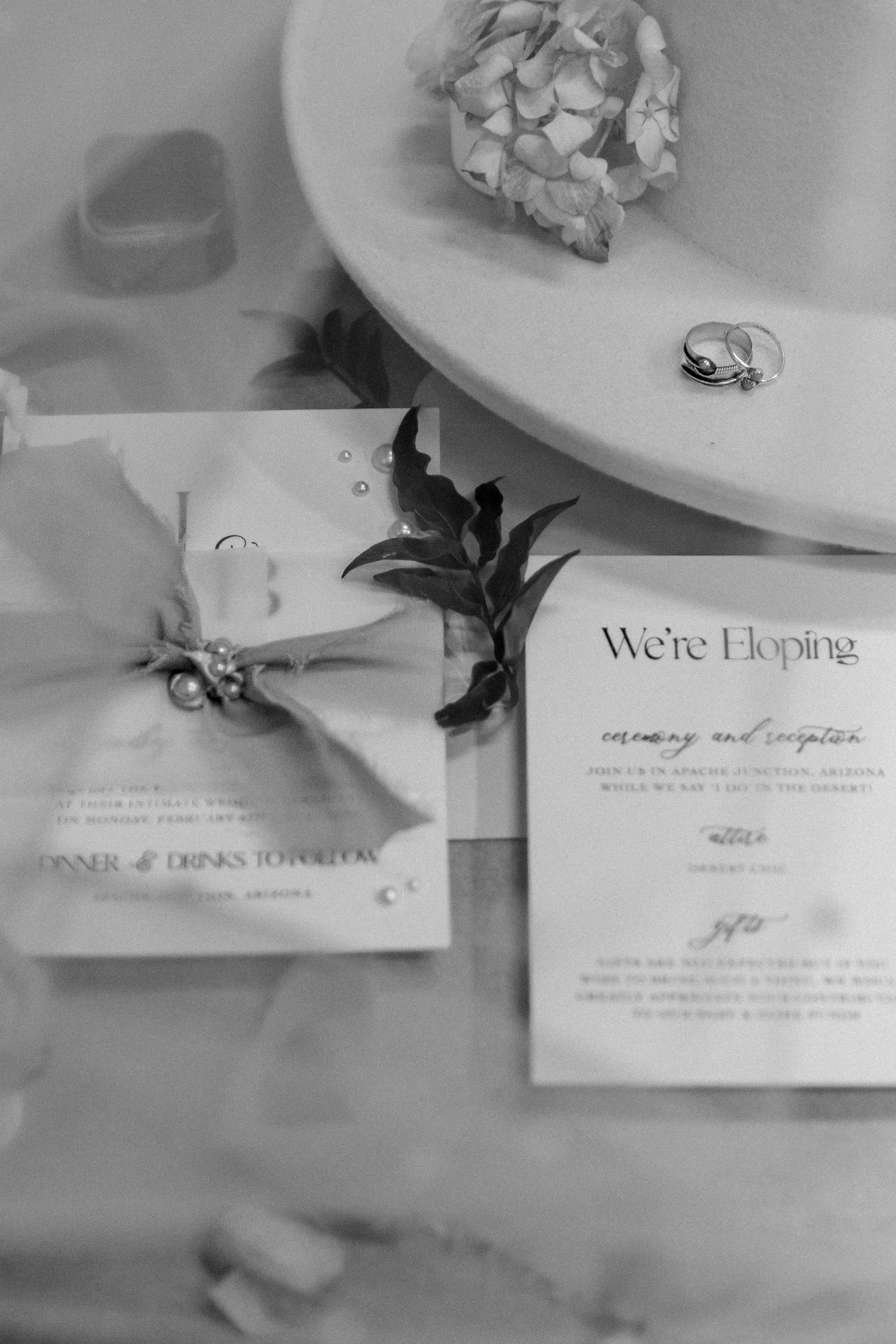 Black and white photo of elopement invitation cards, rings on a hat, flower, and leaves.