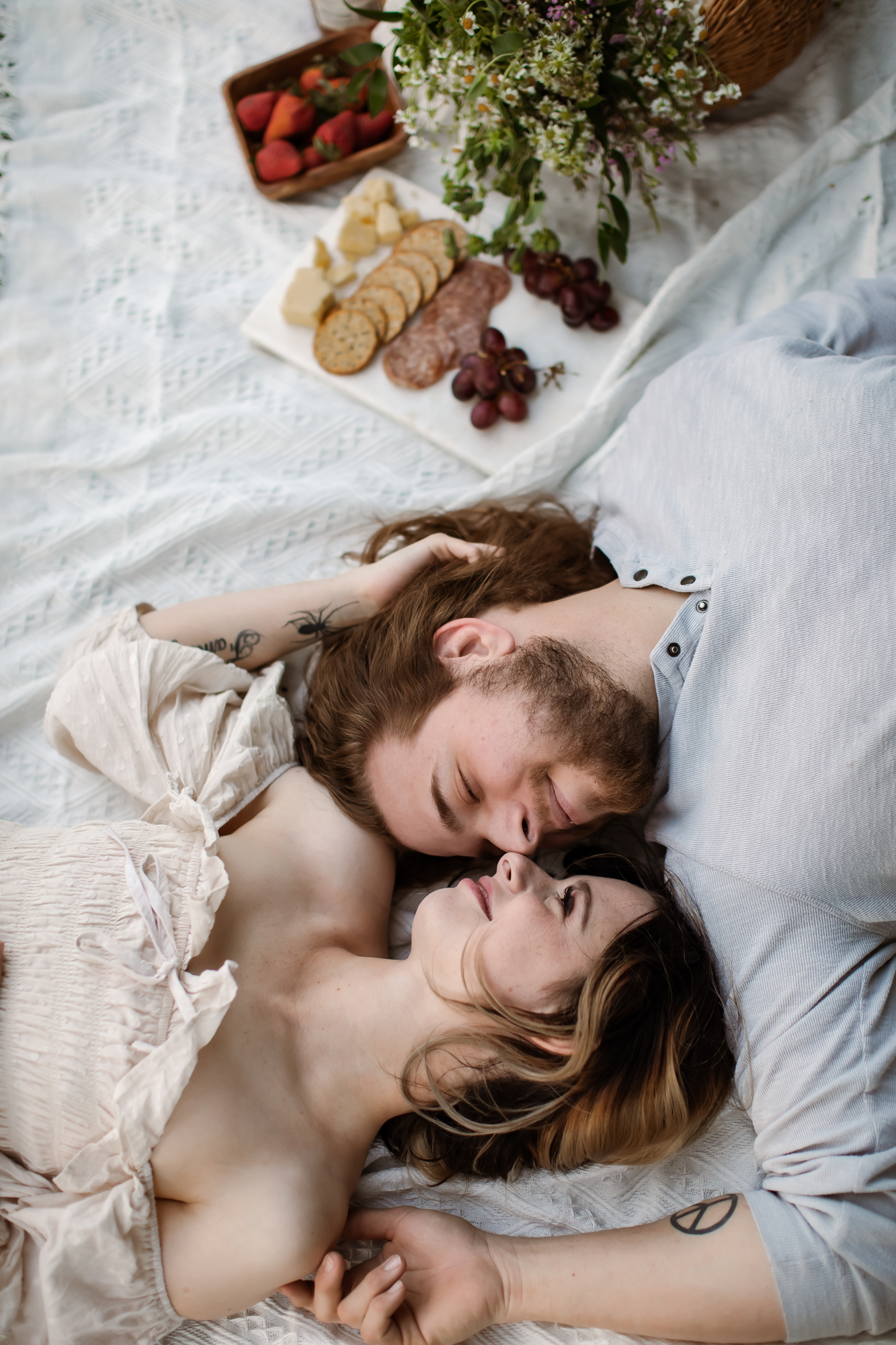 Couple lying on a blanket, gazing into each other's eyes near a picnic with cheese and fruit.