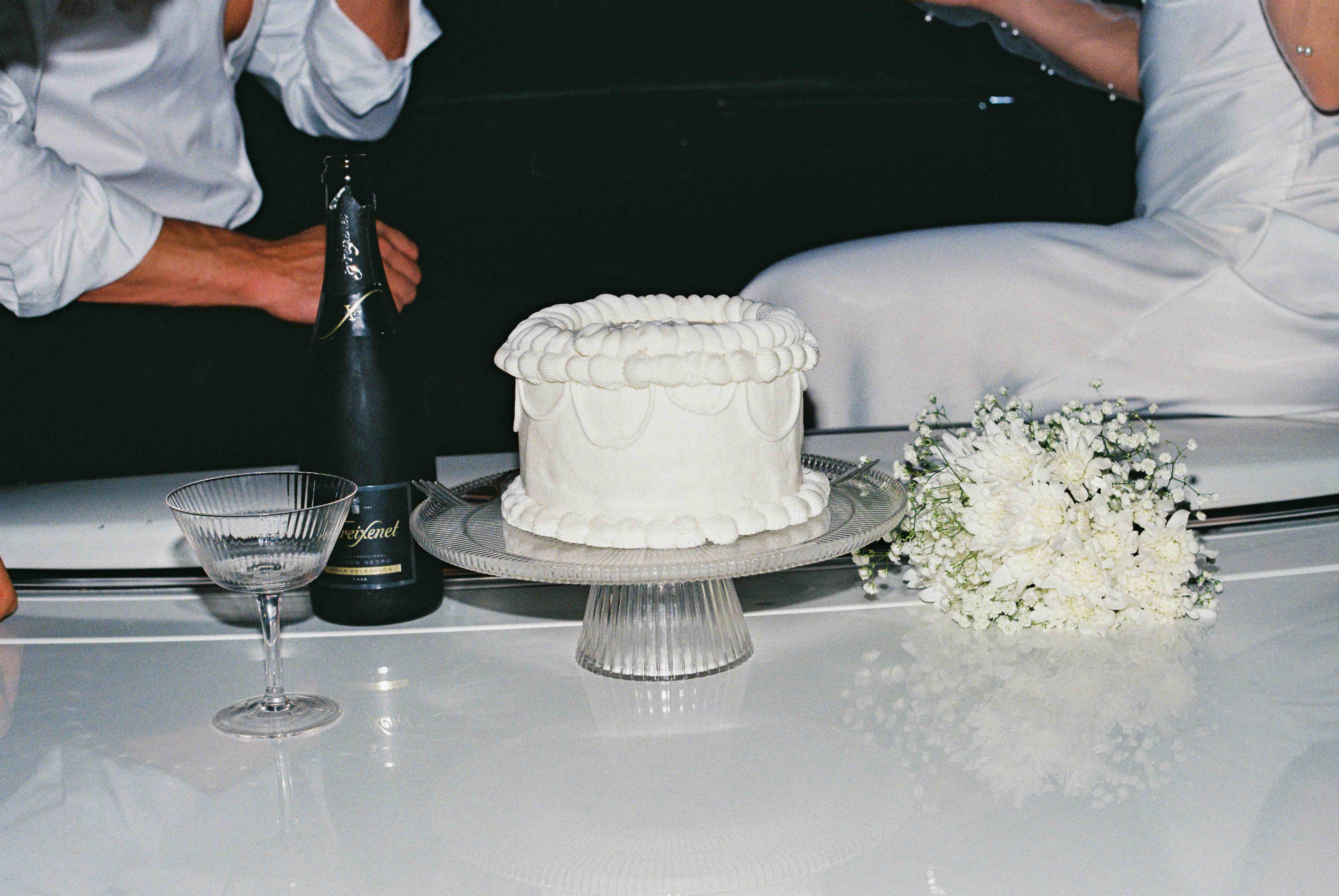 Small white cake on glass stand with white flower bouquet and soda bottle on white surface.