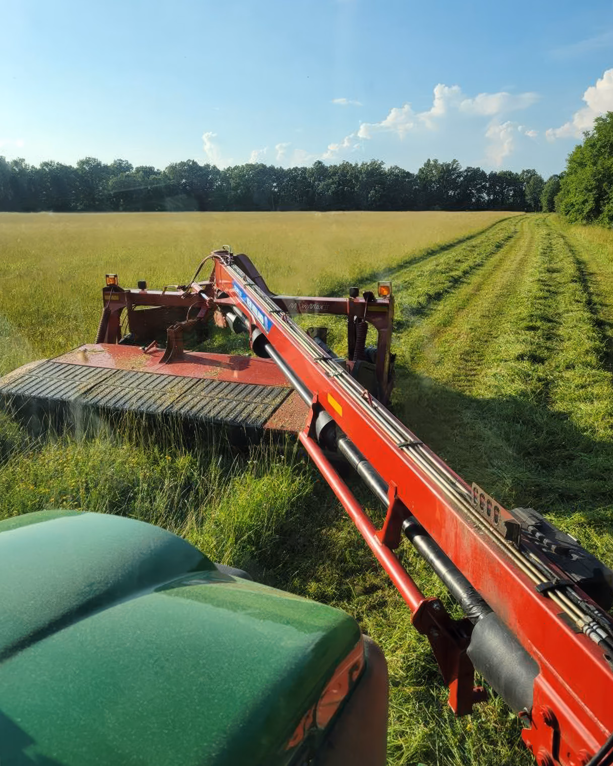 View from tractor cutting grass in a large field with trees lining the horizon under blue sky.