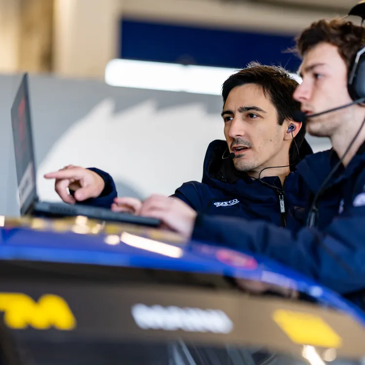 Two men wearing headsets and jackets working on a laptop that's resting on top of a race car, focused and engaged in discussion about weather data.