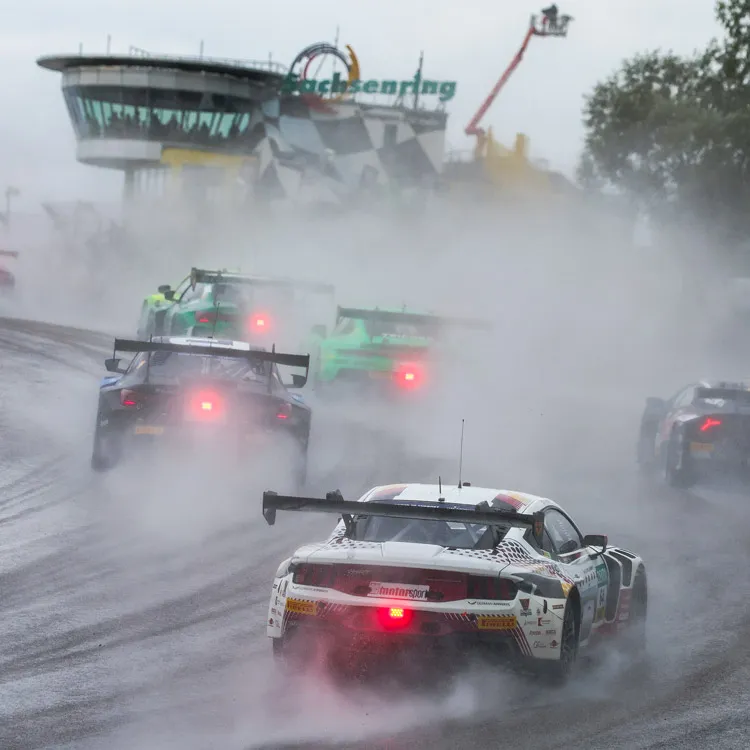 Race cars driving on a wet track creating spray with a control tower visible in the background.