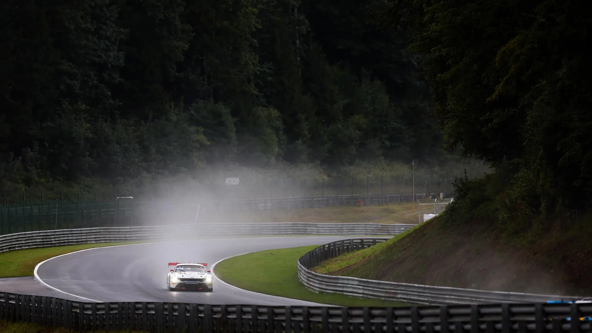 White race car with headlights on driving on wet curved race track surrounded by forest.