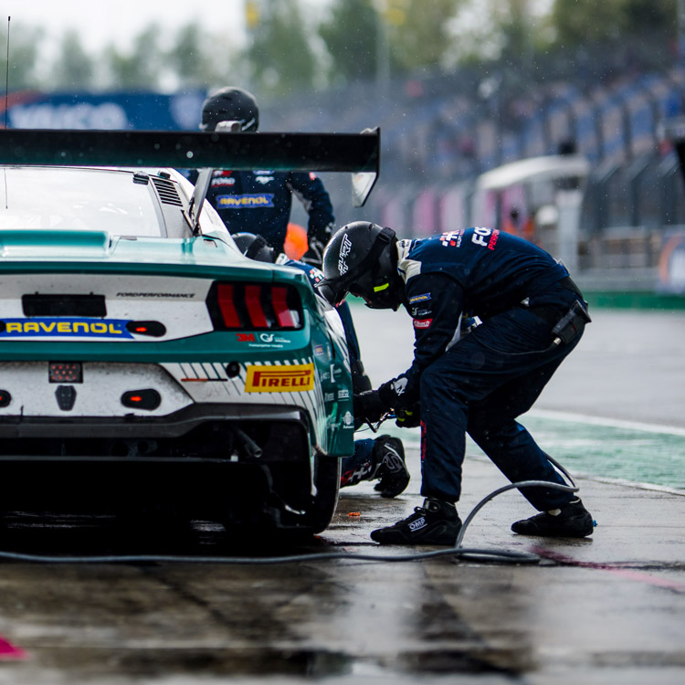 Pit crew members in navy suits and helmets servicing a green and white race car on a wet racing track.