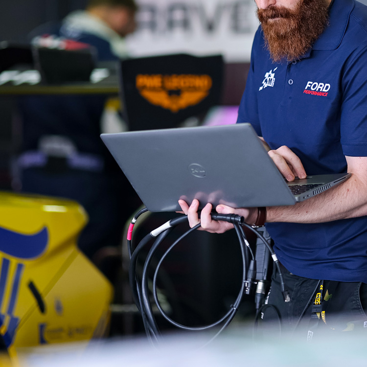 Man in Ford Performance shirt holding a laptop, with a blurred yellow race car in the background.