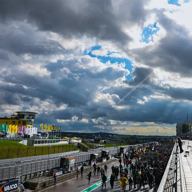 Crowd gathered on a race track pit lane under a dramatic cloudy sky with patches of blue and sunlight.