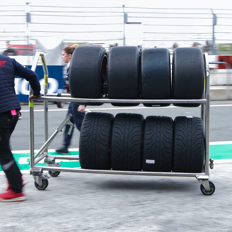 Cart with four slick racing tires on the top rack and four treaded tires on the bottom rack being pushed in a racing pit lane.