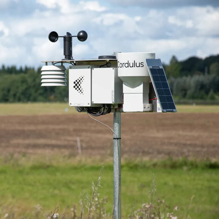 Cordulus weather station with anemometer, solar panel, and rain gauge mounted on a metal pole in a rural field.